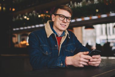 Yarım uzunluk portre denim ceket şık kahve dükkanında dinlenme 4g internet bağlantısı ile Smartphone cep telefonu ile sosyal ağlarda profil güncelleştirilirken kameraya gülümseyen başarılı erkek Blogger