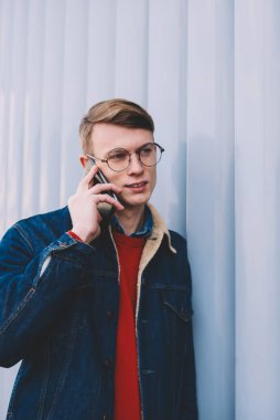 Smartphone üzerinde arama ve mobil faydaları işleci ile görüşmesi sırasında tartışırken rahat giyimli genç adam. Telefon ayakta duvarının yakınında kentsel ortamda konuşurken kot ceketli hipster çocuk