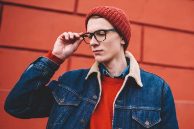 Genç adam şık kırmızı şapka ve optik gözlük vizyon düzeltme aşağı bakıyor. Casual giyinen hipster adam denim ceket ayakta promosyon arka plan üzerinde kentsel ortamda