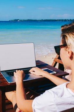 Genç kadının sahte ekran, kadın blogger izlerken videoda netbook dizüstü bilgisayarınızla ve takipçileri ile hafta sonları sohbet yazarak güzel okyanus plaj mesafe çalışmalarına arkadan görünüm