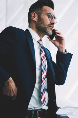 Kendine güvenen orta yaşlı Finans Müdürü smartphone üzerinde arama ve iş ortağı ile konuşurken formal giyim içinde. Gurur CEO'su pencereden dışarı bakarken telefon aygıtı üzerinde iletişim deneyimli