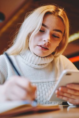 Zeki kız öğrenci ev ödevi yazma üzerine yoğunlaştı. Cep telefonuyla blog içeriğini güncelleyen makale yayınlama fikrine dikkat çeken konsantre kadın blogcu internet haberlerini okumak için cep telefonunu tutuyor.