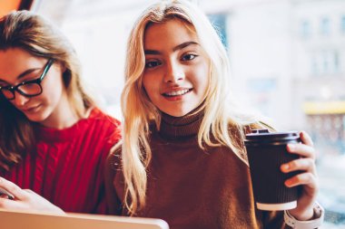 Kahve fincanı tutan neşeli bir kızın portresi kameraya bakıyor arkadaşıyla cep telefonunda sohbet ediyor, serbest çalışan kadınlar kafenin içinde uzaktan çalışmak için teknolojiyi kullanıyorlar.