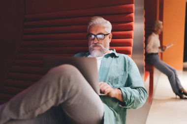 Beyaz sakallı kıdemli beyaz adam dikkatle dizüstü bilgisayar kullanıyor, arka planda odaklanmış bir kadın, canlı kırmızı ve turuncu bir çalışma alanında..