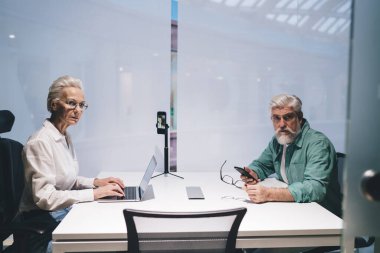 Üst düzey iş arkadaşları ofiste birlikte çalışıyorlar, dizüstü bilgisayarlar ve telefonlar üzerinden projeyi tartışıyorlar. Kadın laptopta yazarken erkek odaklanmış, gözlük ve telefon tutuyor. Verimli bir konuşma.