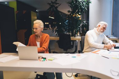 Modern ofiste iş birliği yapan kıdemli meslektaşlarım. Beyaz kazaklı adam notlar alırken kadın belgeleri gözden geçirir. Yeşil bitkiler ve renkli dekorlar yaratıcı çalışma alanını geliştirir