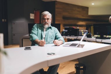 Beyaz saçlı, sakallı, modern ofis masasında dizüstü bilgisayarı ve akıllı telefonuyla oturan kıdemli beyaz iş adamı. İşten ya da tefekküre ara vermek. Sıcak bir ofis ortamı.