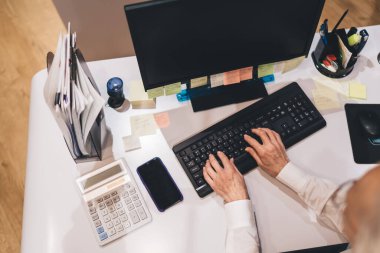 Düzensiz masadaki klavyeye yazı yazan kişilerin genel görünümü yapışkan notlar, hesap makinesi, dokümanlar ile birlikte izleniyor.Düzenli bir çalışma ortamında muhasebe veya finansal görevler üzerine çalışan