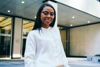 Başarılı Afrikalı-Amerikalı kadın ofis çalışanı portresi finans belgeler klasörüyle tutarak ve kentsel belirleyen ve kameraya gülümseyen ayakta kamera gülümseyen beyaz gömlek giymiş