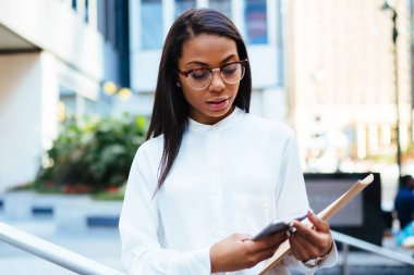 Elinde belgeler klasörüyle tutan smartphone aygıt üstünde posta kontrol dalgın Afrikalı-Amerikalı kadın memur. Koyu tenli çalışan hücresel ayakta açık havada sokağa haber okuma