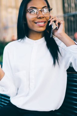 Cep telefonu arama ve şehir merkezindeki bankta oturan konuşma sırasında gülüyor trendy gömlekli neşeli koyu tenli kadın ofis çalışanı. Pozitif genç kadın smartphone aygıt üzerinde söz