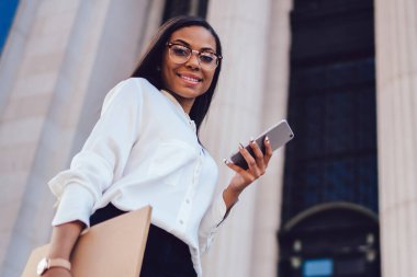 Aşağıda neşeli Afrikalı-Amerikalı kadın çalışanın görünümü smartphone ücretsiz 4 g internet ayakta mimari building yakınındaki kentsel ortamda üzerinden mobil uygulaması güncelleştiriliyor beyaz gömlek giymiş