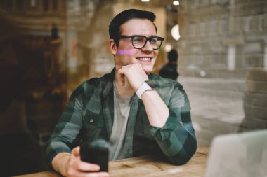 Neşeli hipster adam uzakta smartphone, müreffeh erkek freelancer mesafesi iş modern aygıtlar ve kablosuz internet ile kafeterya iç memnun tutan penceresinde arıyorum gözlükler