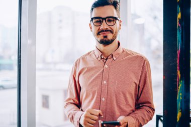Sıradan giyinen, akıllı telefon takan, kameraya bakan, sosyal ağlarda sohbet etmek için telefonu kullanan, gözlüklü, yakışıklı hippi bir genç girişimcinin yarım boy portresi.