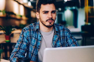 Üniversitede internet ve kablosuz internet kullanarak sınav hazırlığı yapan ciddi bir hipster çocuğun portresi. Kameraya bakan genç adam, birlikte çalıştığı yerdeki dizüstü bilgisayarları kullanarak hayal kırıklığına uğradı.