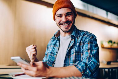 Şapkalı neşeli hippi adamın portresi çevrimiçi yarışmada kazandığı zaferle sevinçten havalara uçuyor telefonu elinde tutarak sınavlardaki başarılarını kutlayan genç adam başarılı bir şekilde mesaj alıyor.