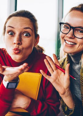 Yarım uzunluk portre pozitif öğrencinin hava öpücük ve güzel sarışın hipster blogger birlikte rekreasyon vaktinizi el sallayarak gözlük gönderme. Mutlu genç kadın eğlenceli kameraya gülümseyen