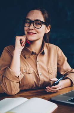 Yarım uzunluk portre ise smartphone elinde tutan ve uzaktan kablosuz internet'e bağlı dijital dizüstü bilgisayarda çalışan kameraya gülümseyen gözlük mutlu kadın grafik tasarımcısının