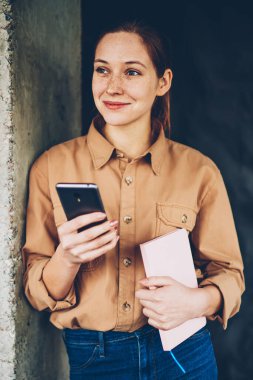 El smartphone aygıt yolu ile yüksek hızlı 4 g internet ayakta kopyalama alanı alanı yakınındaki posta kontrol kitap ile gülümseyen düşünceli genç kadın. Neşeli kadın blogger ile modern cep durulması