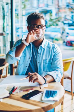 Yakışıklı akıllı yetenekli erkek mimar trendy gözlük uzakta kafeteryasında cam fincan içme suyu seyir içinde. Olgun zeki hindu çalışan çalışma sonu tahta masada oturan zevk
