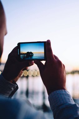 Gün batımında sakin deniz suyunun video kaydını tutan bir adamın kırpılmış görüntüsü. Akşam karanlığında erkek, iyi kalite kamera ve akıllı telefon uygulaması kullanarak doğanın fotoğrafını çekiyor.