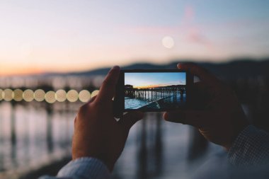 Doğanın akşam karanlığında modern akıllı telefon kamerasıyla fotoğraf çeken bir adamın kırpılmış görüntüsü, cep telefonu tutan hipster bir adamın Alacakaranlık 'ta deniz suyu uygulaması ile fotoğraf çekerken görüntüsü.