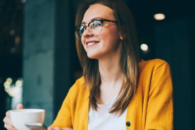 Gözlüklü, gözlüklü, elinde akıllı telefon aleti ve elinde fincanı olan mutlu bir kadın. Kafeteryada iletişim kurmak için cep telefonu bağlantısını kullanırken yüzünü çeviren pozitif hippi bir kız.