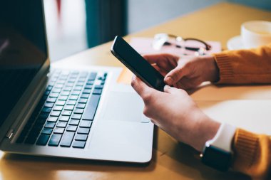 İyi internet bağlantısı olan akıllı telefonları olan, kafeteryada dizüstü bilgisayarla oturan, cep telefonuyla seyahat planları yapan bir kadının kesilen görüntüsü.