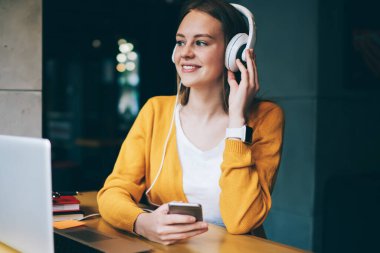 Kulaklıklı neşeli bayan öğrenci dizüstü bilgisayardan öğrendikten sonra müzik molası veriyor, genç pozitif hipster kız akıllı telefon cihazında multimedya uygulaması ile ses kayıtlarını dinliyor.