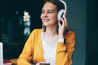 Kulaklıklı neşeli bayan öğrenci dizüstü bilgisayardan öğrendikten sonra müzik molası veriyor, genç pozitif hipster kız akıllı telefon cihazında multimedya uygulaması ile ses kayıtlarını dinliyor.