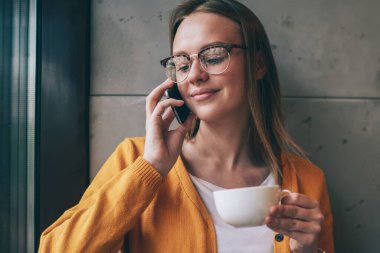 Gözlüklü pozitif bayan öğrenci göz koruması sağlıyor. Kahve molası sırasında arkadaşça telefon konuşmalarının tadını çıkarıyor. Kapalı mekanda, elinde sıcak çay bardağıyla mutlu, çekici hippi kız.
