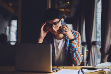 Şık gözlüklü genç serbest çalışan, kafenin içinde otururken akıllı telefondan konuşan ve modern laptop cihazında çalışan bir öğrenci. Ödev yaparken cep telefonuyla iletişim kuran bir öğrenci.