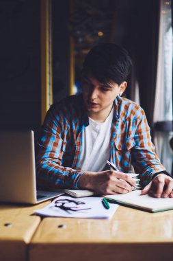 Zeki hippi, dizüstü bilgisayara bakıyor ve not defterine bilgi yazıyor. Kafede oturmuş internetten ilginç bir Webinar izlerken not alan dalgın genç adam.