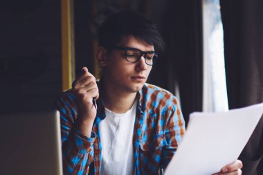 Zeki öğrenci kağıt üzerinde yazılı metin bilgilerini okuyor. Diz üstü bilgisayar ve kitaplarla şık bir kahve dükkanında oturuyor. Hipster adam elinde kalem tutuyor ve derslere bakıyor.