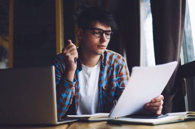 Zeki öğrenci kağıt üzerinde yazılı metin bilgilerini okuyor. Diz üstü bilgisayar ve kitaplarla şık bir kahve dükkanında oturuyor. Hipster adam elinde kalem tutuyor ve derslere bakıyor.