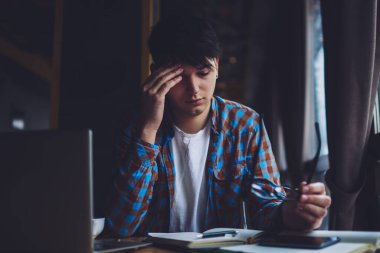 Elde gözlüklü, optik gözlüklü, kitap okuyan ve sınavlara hazırlanan yorgun genç bir adam modern bilgisayar cihazıyla birlikte çalışıyor. Ödev yapmaktan yorgun düşmüş bir öğrenci.