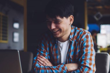 Neşeli hippi çocuk dizüstü bilgisayardan komik videolar izlerken gülüyor. Mutlu öğrenci internet sitelerinde pozitif hikaye okuyor. Kafenin içindeki ahşap masada oturan modern bilgisayarı kullanıyor.