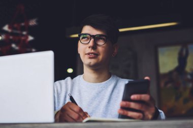 ?Gözlüklü, elinde akıllı telefonla not defterine mesaj yazan yakışıklı bir adam. Ev ödevi hakkında düşünen düşünceli bir öğrenci dizüstü bilgisayarın başında oturup bazı bilgileri not alıyor.