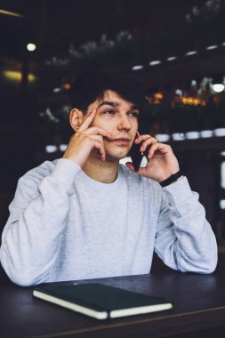 Modern akıllı telefondan konuşan ve çözümü düşünen meşgul genç girişimci. Kafede ders kitabıyla ahşap masada oturan düşünceli öğrenci ciddi cep telefonu konuşmaları yapıyor.