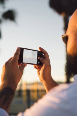 Erkek blogger fotoğraf çekmek için sahte ekranı ile smartphone kullanarak görünümünü sırt ve ağlarda, modern cep telefonu fotoğrafları yaz tatillerini akşamları sırasında yapma hipster adam pay