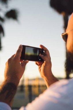 Dikiz genç erkek tutan eller smartphone ile modern fotoğraf makinesi için güzel gün batımı, fotoğraflarını alarak kırpılmış hipster adam cep telefonu üzerinde akşam manzara çekim görünümünü yakın