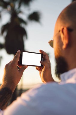 Erkek blogger fotoğraf çekmek için sahte ekranı ile smartphone kullanarak görünümünü sırt ve ağlarda, modern cep telefonu fotoğrafları yaz tatillerini akşamları sırasında yapma hipster adam pay