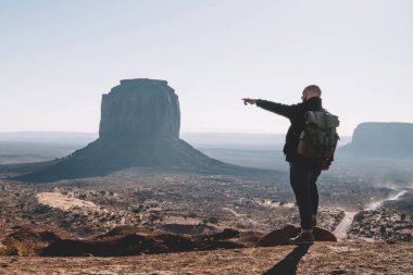 Güneybatı, turistik Yolculuk tutkusu manzarayı kuru çöl kumtaşı oluşumu ile sırt çantası içinde ABD hiking ile ünlü anıt Vadisi yüksek kayalar sahne görünümü işaret eden erkek Gezgin