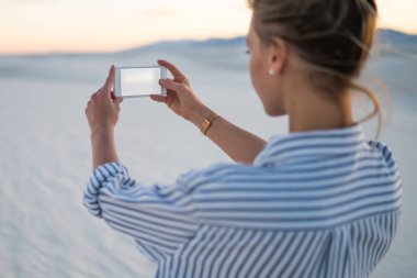 Genç kadın turist beyaz kum tepeleri, keşfetmek smartphone kamera Hipster kız Yolculuk tutkusu günbatımı sahne görünümünü tatil zevk cep telefonu kamera fotoğraf çekimi selfie yapma arkadan görünüm