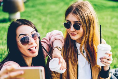 Mutlu genç hippi kız birlikte modern smartphone kamerada fotoğraf yapma dinlenme zevk. Mobil sosyal ağ Park yeşil çimenlerin üzerinde oturan paylaşımı için selfie için poz neşeli arkadaşlarım