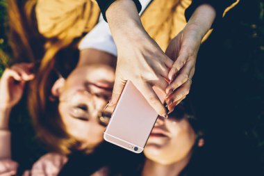 Kadının el dijital smartphone ve fotoğraf çekimi arkadaşıyla çim üzerinde yalan özel fotoğraf uygulaması üzerinde tutarak en iyi seçim odak. Selfie yapma iki kadın blogcular arka plan bulanık