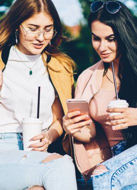 Esmer blogger posta lezzetli kahve zevk bankta oturan modern telefon aygıtı üzerinde en iyi arkadaşı için Internet Web sitesinde gösterilen. Hippi kız giyim fotoğraflarını smartphone açık havada üzerinde tartışıyor
