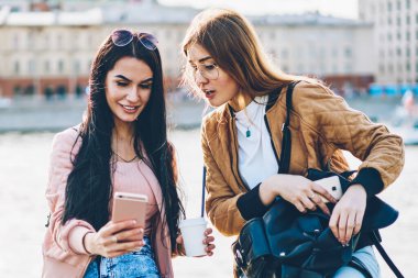 İki neşeli arkadaşlarım şık siyah sırt çantası ve lezzetli kahve el modern telefon şehir promosyon arka plan üzerinde oturan yüklü yeni fotoğraf uygulaması ile selfie resim yapma