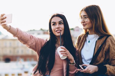 İki güzel gezginler yapma selfies fotoğraflar üzerinde modern telefon ayakta zekâ kahve ve sırt çantası şehir promosyon arka plan üzerinde reklam için reklam alanı yakınında şık casual kıyafet ile