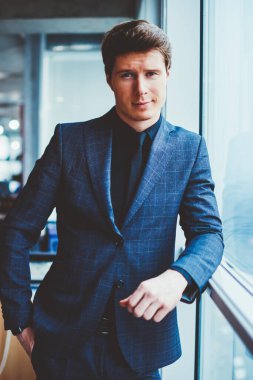 Kendine güvenen şık takım elbiseli bir işadamının portresi ofis penceresinin yanında duruyor ve kameraya bakıyor, yakışıklı beyaz erkek girişimci çalışma alanında vakit geçiriyor ve iş arkadaşını bekliyor.